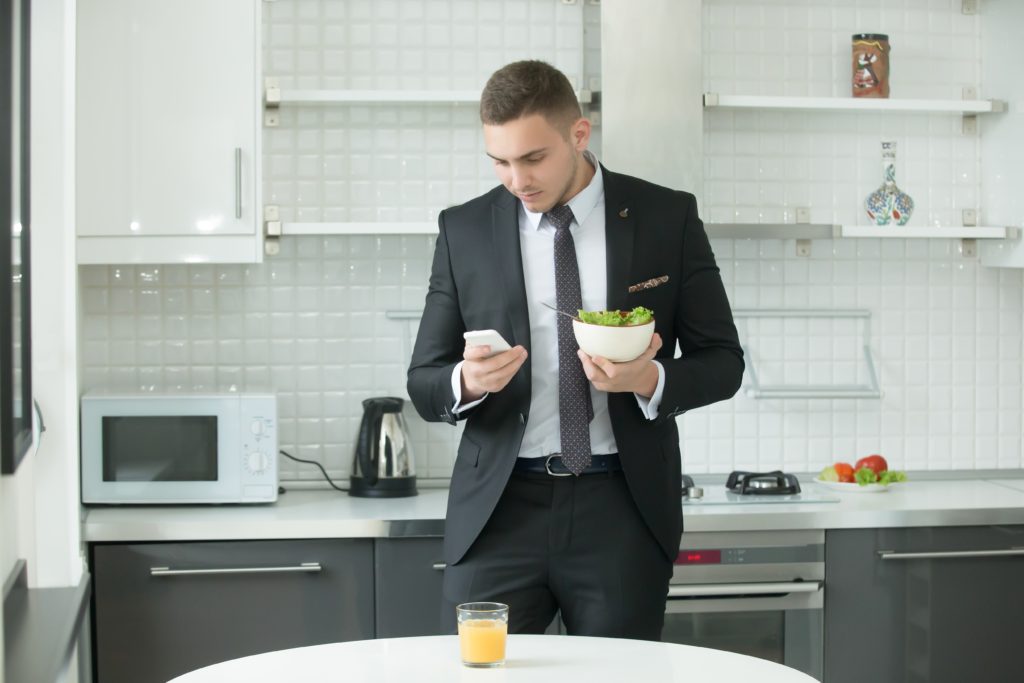 Young handsome businessman at the modern kitchen having vegan lunch, looking at his smartphone. Office worker having breakfast before working day at home