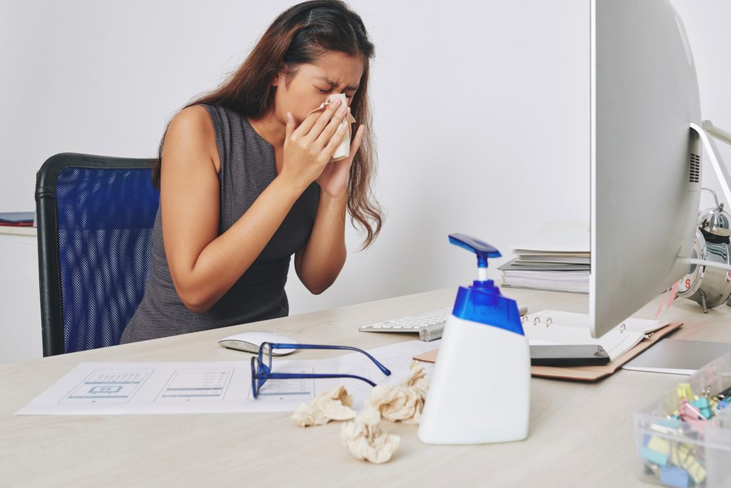 Young businesswoman sneezing in paper tissue when sitting at her desk, flu and allergy concept