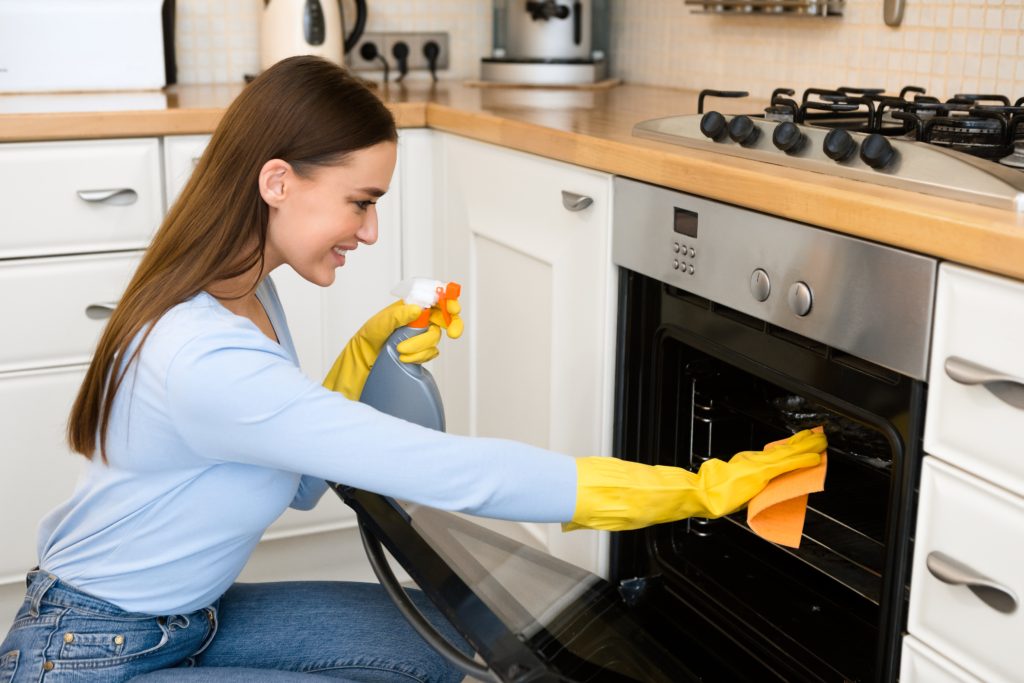 Kitchen Chores Concept. Girl in yellow protective rubber gloves cleaning dirty oven with microfiber rag and spray